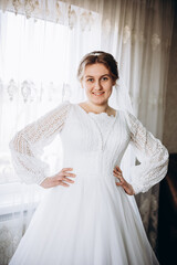 A beautiful bride in a white lace wedding dress smiles softly indoors. She stands by a window with sheer curtains, looking radiant and happy on her special day before the ceremony.