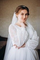 A beautiful bride in a white lace wedding dress smiles softly indoors. She stands by a window with sheer curtains, looking radiant and happy on her special day before the ceremony.