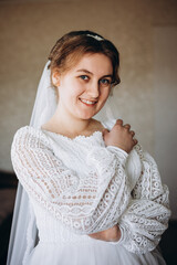 A beautiful bride in a white lace wedding dress smiles softly indoors. She stands by a window with sheer curtains, looking radiant and happy on her special day before the ceremony.