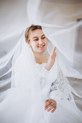 A smiling bride in a white lace wedding dress poses with her veil flowing gracefully around her. The soft fabric and natural light create a romantic and dreamy bridal portrait.