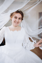 A smiling bride in a white lace wedding dress poses with her veil flowing gracefully around her. The soft fabric and natural light create a romantic and dreamy bridal portrait.