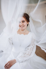 A smiling bride in a white lace wedding dress poses with her veil flowing gracefully around her. The soft fabric and natural light create a romantic and dreamy bridal portrait.