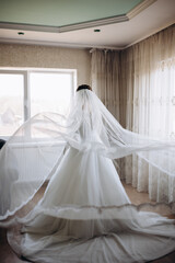 Bride in a luxurious white dress stands by the window, her veil gently flowing in the air. A scene filled with tenderness, light, and anticipation of a special wedding moment.