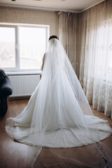Bride in a luxurious white dress stands by the window, her veil gently flowing in the air. A scene filled with tenderness, light, and anticipation of a special wedding moment.