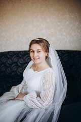 A beautiful bride in a white lace wedding dress smiles softly indoors. She stands by a window with sheer curtains, looking radiant and happy on her special day before the ceremony.