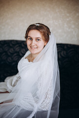 A beautiful bride in a white lace wedding dress smiles softly indoors. She stands by a window with sheer curtains, looking radiant and happy on her special day before the ceremony.
