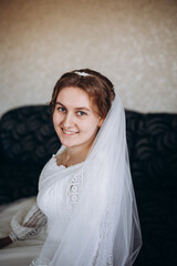 A beautiful bride in a white lace wedding dress smiles softly indoors. She stands by a window with sheer curtains, looking radiant and happy on her special day before the ceremony.