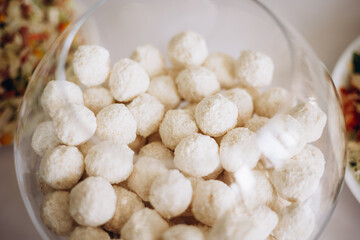 A glass bowl filled with coconut candies shaped like white balls. Light desserts coated in coconut flakes create a tender and appetizing look on the festive table.