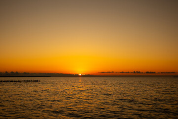 Sunset over calm sea with orange sky, distant clouds on horizon and visible breakwater.