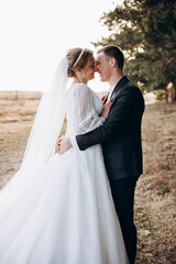 The newlywed couple walks along a natural path, holding hands. The bride in a long white dress looks back with a smile while the groom walks beside her. A warm and peaceful outdoor moment.