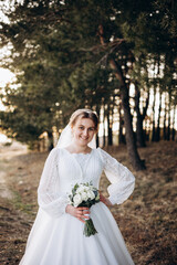 A bride in a beautiful white lace dress stands outdoors holding a bouquet of white roses. A calm and tender atmosphere of a wedding day in nature.