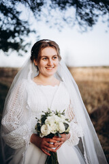 A bride in a beautiful white lace dress stands outdoors holding a bouquet of white roses. A calm and tender atmosphere of a wedding day in nature.