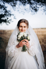 A bride in a beautiful white lace dress stands outdoors holding a bouquet of white roses. A calm and tender atmosphere of a wedding day in nature.