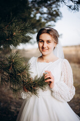 Bride in a white lace wedding dress posing outdoors near pine branches. Natural makeup and soft smile. Rustic countryside wedding portrait in warm daylight.