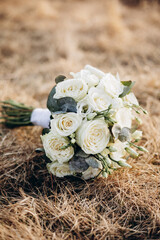 Elegant bridal bouquet with white roses and eucalyptus lying on dry grass. Romantic wedding still life with soft natural light and rustic background