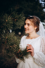 Bride in a white lace wedding dress posing outdoors near pine branches. Natural makeup and soft smile. Rustic countryside wedding portrait in warm daylight.