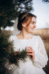 Bride in a white lace wedding dress posing outdoors near pine branches. Natural makeup and soft smile. Rustic countryside wedding portrait in warm daylight.