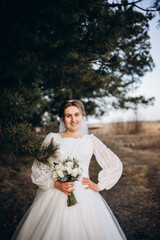 Bride in a white lace wedding dress posing outdoors near pine branches. Natural makeup and soft smile. Rustic countryside wedding portrait in warm daylight.