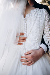 Close-up of a groom gently holding the bride in a white lace wedding dress. Elegant details of buttons and veil. Romantic and tender wedding moment.