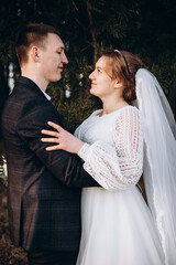 A newlywed couple poses in a forest, embracing warmly. The bride wears an elegant white dress and holds a bouquet of white flowers while the groom smiles beside her. A tender moment captured in nature