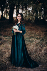 A woman in a long emerald velvet dress stands outdoors holding a bouquet of white flowers. She poses calmly, creating an elegant and gentle portrait in a natural forest setting. The bridesmaid 