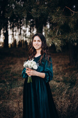 A woman in a long emerald velvet dress stands outdoors holding a bouquet of white flowers. She poses calmly, creating an elegant and gentle portrait in a natural forest setting. The bridesmaid 
