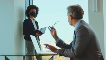Business executive and professional assistant discussing work during a meeting in a modern corporate office, both wearing protective face masks for health safety