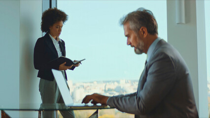 Business professionals collaborate in a modern office, woman reviews planner while colleague types on laptop by a bright window, focused on planning and teamwork