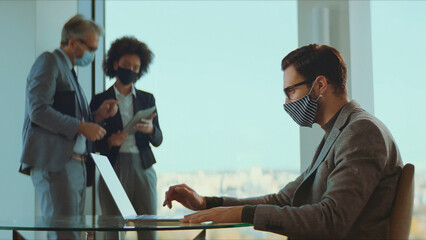 Business colleagues in masks work in a modern office: one man types on a laptop while two coworkers discuss strategy in the background, practicing pandemic safety and teamwork