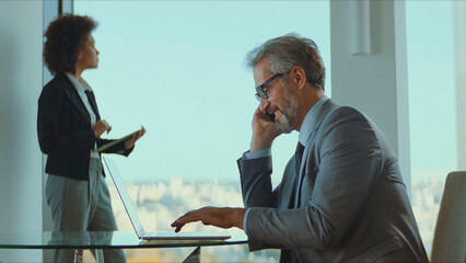 Business executive talking on a mobile phone and typing on a laptop with a female colleague standing in the background, both professionally dressed in a modern office with panoramic city views