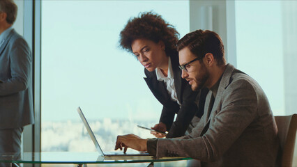 Business professionals engaging in a focused discussion while working collaboratively on a laptop at a glass desk during a corporate meeting in a bright high-rise office