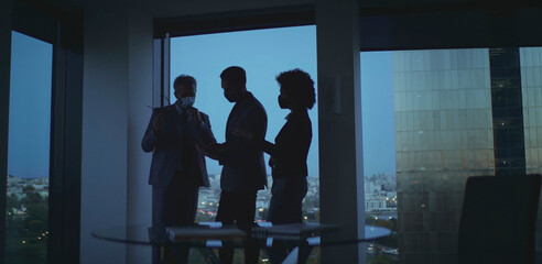 Business professionals in suits wearing face masks stand by a large office window at dusk, overlooking a city skyline while holding a corporate meeting and planning strategy