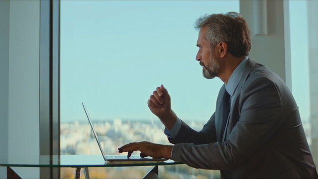 Experienced senior corporate leader diligently typing on a laptop, reflecting expertise and professionalism while staying connected in a bright contemporary office with a city view