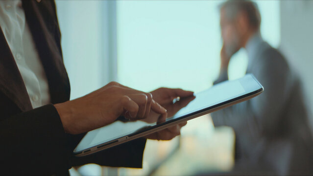 Business executive hands operating a modern digital tablet, interacting with technology during a corporate meeting with a colleague in the background, focusing on data and networking