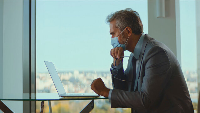 Mature businessman wearing a protective face mask while working on a laptop at a modern office desk against a city skyline, reflecting on business challenges and the new normal