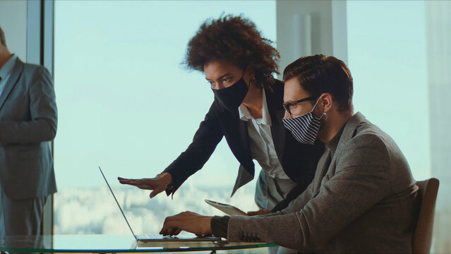 Business colleagues wearing face masks collaborate over a laptop in a modern office, practicing safety and teamwork during the pandemic while planning and discussing strategy