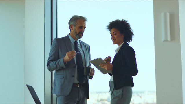 Two business professionals, diverse man and woman, standing in a bright office reviewing a tablet together, demonstrating teamwork, modern communication and collaborative planning