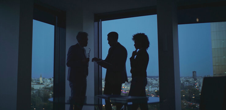 Business team collaborating on strategy during an evening meeting in a modern office with city skyline view, silhouettes of executives planning and discussing a project - Powered by Adobe