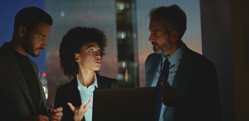 Diverse business colleagues working together, discussing a project on a laptop in a modern office at night, demonstrating teamwork and professional collaboration