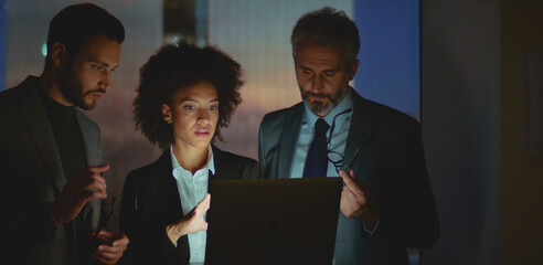 Diverse business professionals working together in a corporate office, a dedicated team collaborating after hours, focusing on a laptop screen to meet deadlines and drive project success