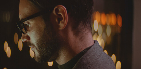 Man with beard and glasses concentrating on a task, illuminated by a screen, with blurred city lights creating bokeh in the background, symbolizing dedication and working extended hours