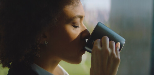 Woman with eyes closed slowly sipping from a dark coffee mug, finding peace and relaxation while standing by a window, bathed in warm morning sunlight
