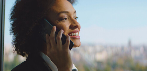Happy businesswoman having a phone call, standing by a large window with a blurry city skyline in...