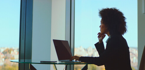 Thoughtful businesswoman looking out of a large office window, contemplating new ideas and future strategies while working on a laptop, with a cityscape providing a background view