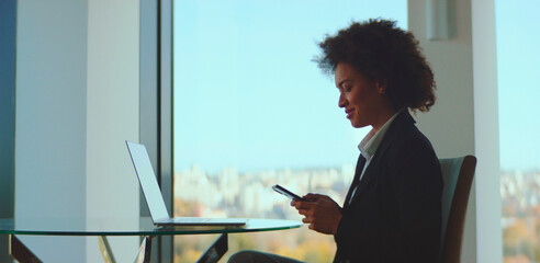 Black businesswoman sitting by an office window, smiling while using smartphone with laptop on glass table and city view, conveying modern professional confidence and connectivity