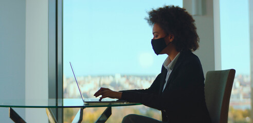 Young black businesswoman wearing protective face mask diligently working on laptop computer during coronavirus pandemic while sitting at desk in modern office with city view background