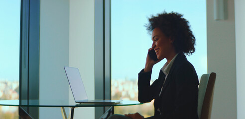Confident businesswoman smiling while having a phone conversation, using a mobile device in a contemporary high-rise office with a laptop on a glass table