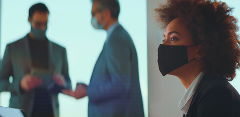 Attentive businesswoman in black protective face mask and professional attire attending a corporate meeting with colleagues, practicing safety and social distancing during pandemic