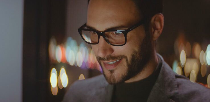 Young man wearing glasses and a beard smiling while working at night, the city lights creating a bokeh effect in the background and a screen reflection in his eyewear