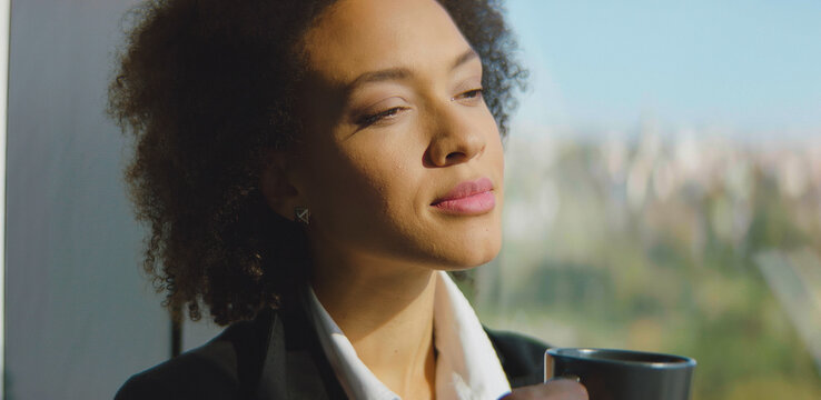 Young african american businesswoman drinking coffee, relaxing herself during a break, looking outside a window with an urban background, thinking about work and having ideas - Powered by Adobe